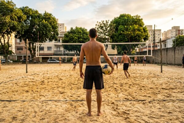 Jogador de costas atrás da linha de fundo da quadra de areia segurando a bola antes do saque